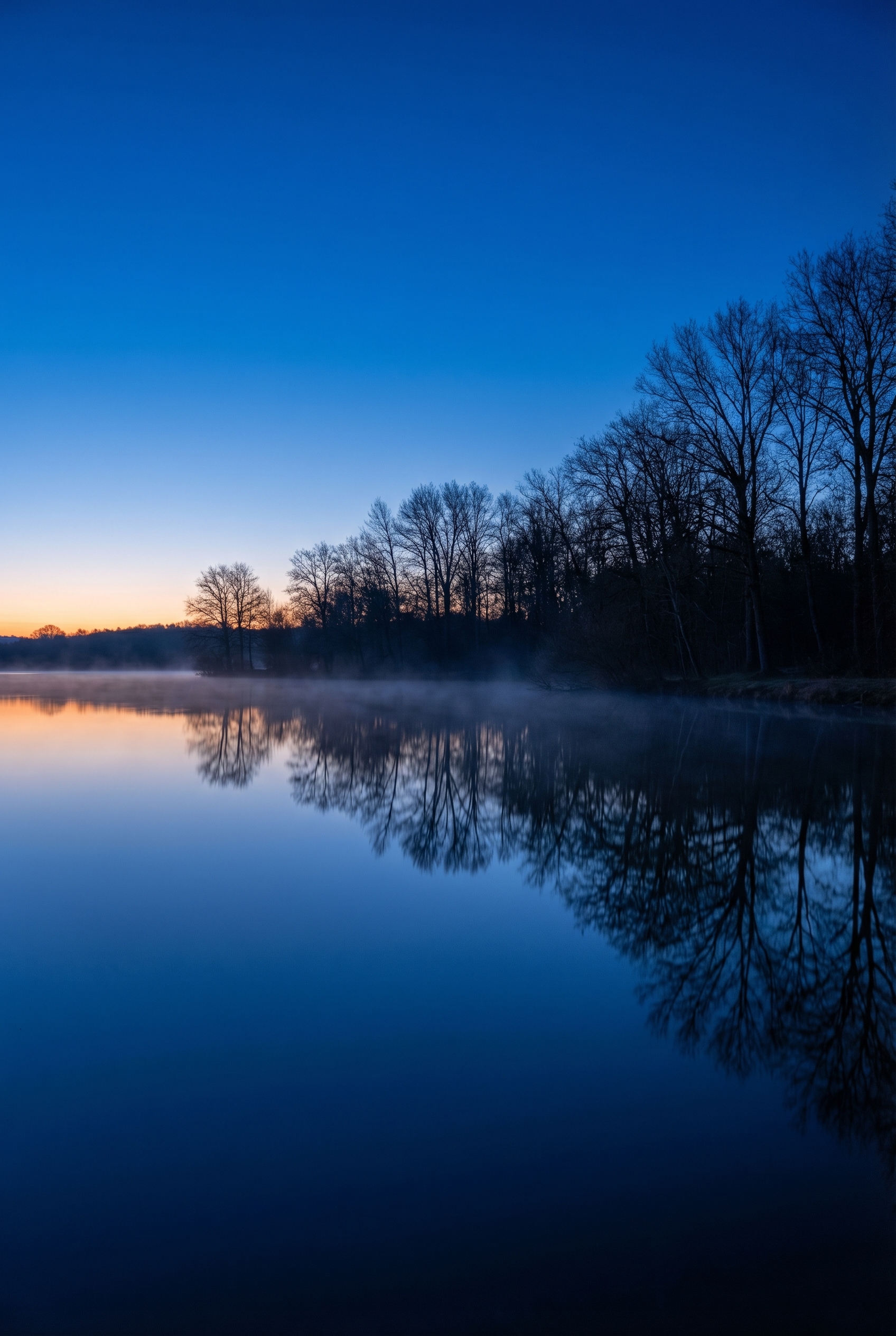 Golden-hour : comment (bien) la photographier 27 Paysage de lac en France à la fin de l'heure dorée avec reflets de silhouettes d'arbres, brume légère sur l'eau et dégradé de ciel orangé et bleu.