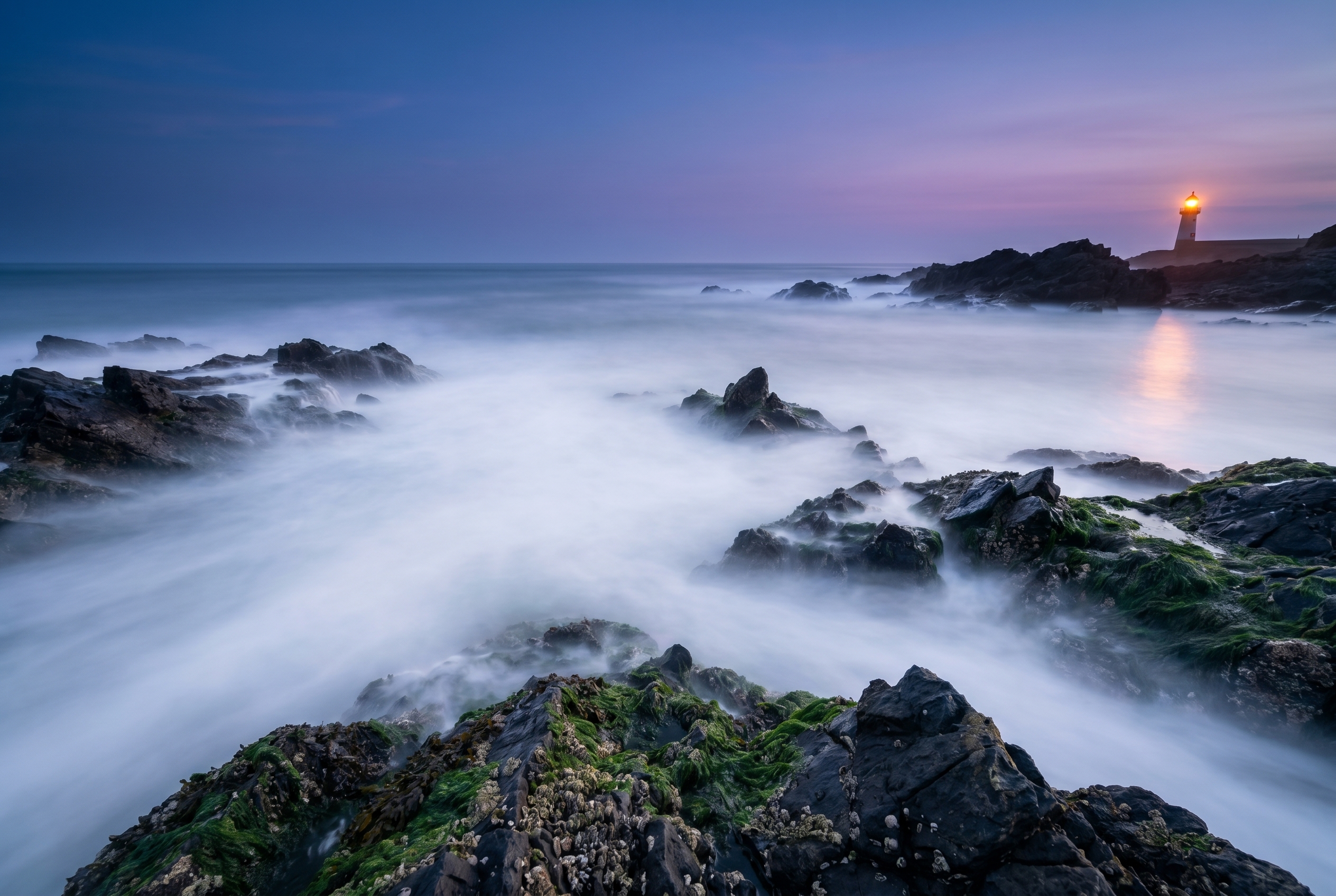 Photographie de paysage marin avec pose longue montrant une mer cotonneuse entourant des rochers côtiers à l'heure bleue.